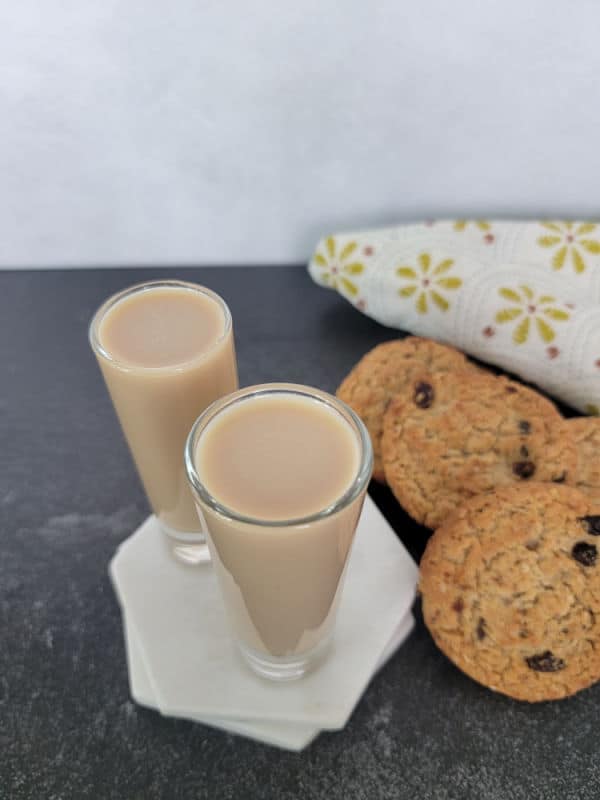 Two cocktail shots on a white marble coaster next to a stack of oatmeal cookies and a cloth napkin 