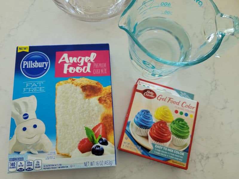 Angel food cake mix, food coloring, and a measuring glass with water on a white counter