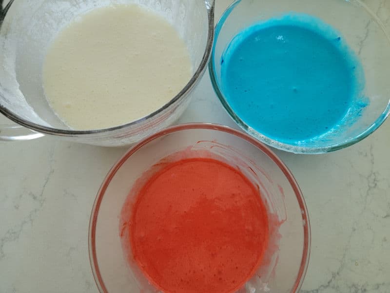 Glass bowls with white, blue, and red batter in them on a white counter