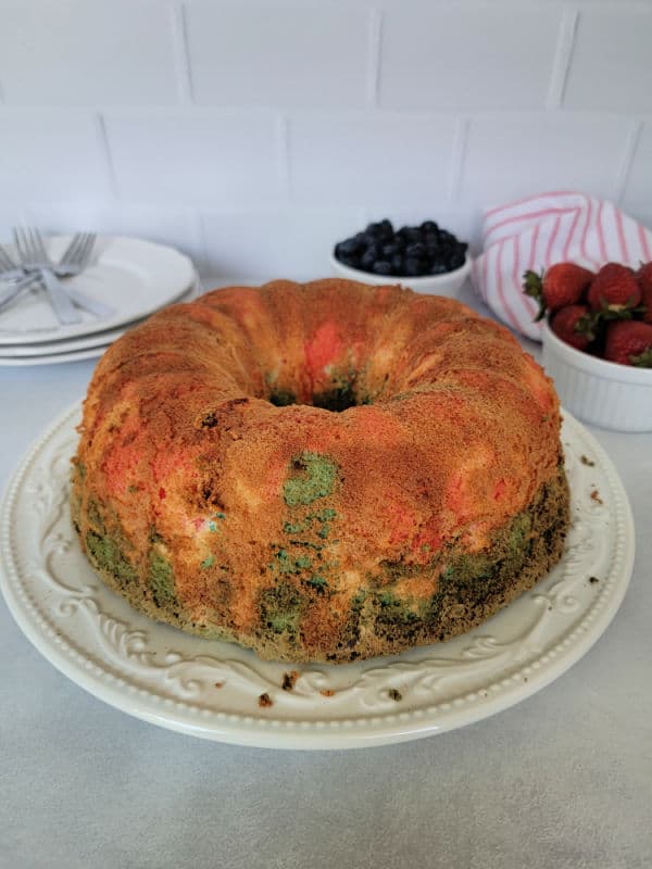 Multi colored Angel food cake on a white plate next to a bowl of blueberries and strawberries, plus a stack of plates with forks. 