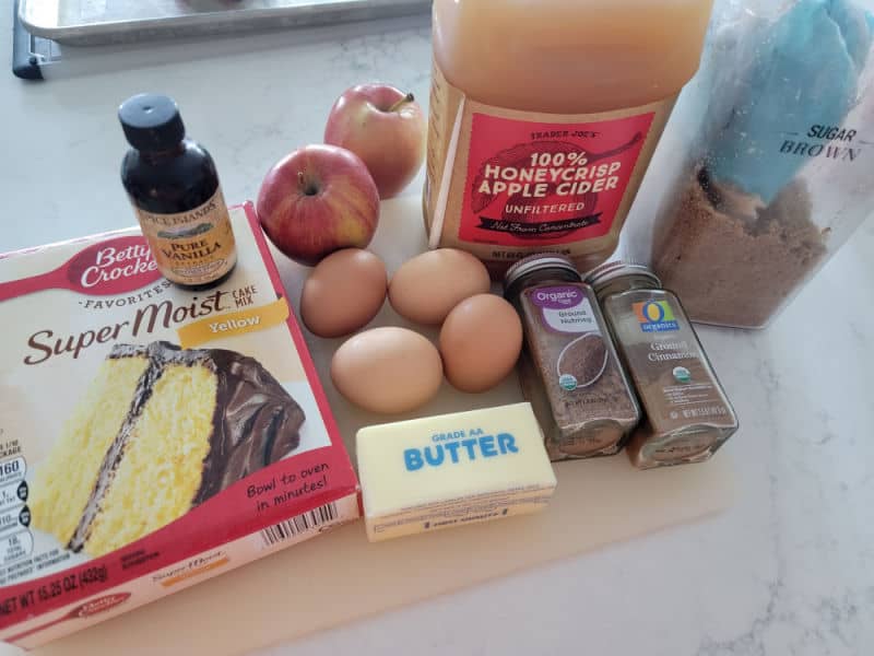 Apple Cider Donut Cake ingredients, yellow cake mix, vanilla, eggs, apples, apple cider, brown sugar, butter, nutmeg, and cinnamon on a cutting board