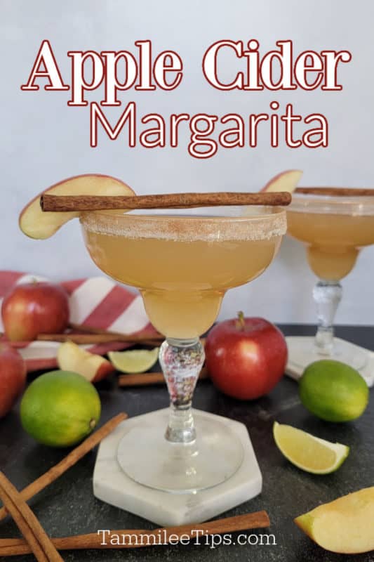 Apple cider margarita over a margarita garnished with an apple slice and cinnamon sticks surrounded by apples, limes, and cinnamon sticks. 