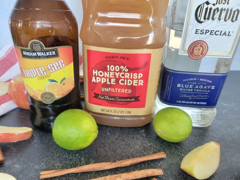 Triple sec, apple cider, Jose Cuervo tequila next to apple slices, cinnamon sticks, and limes 