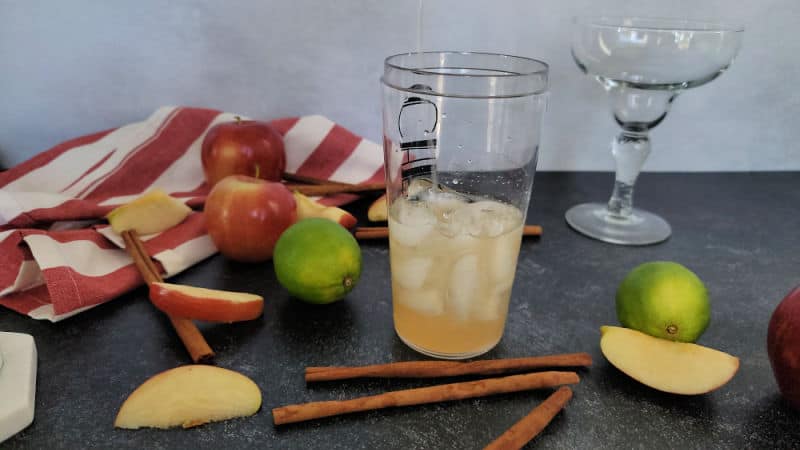 Cocktail shaker with ice and liquid next to a margarita glass, apples, apple slices, cinnamon sticks, and limes