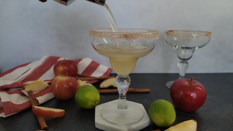Cocktail shaker pouring into a margarita glass next to apples, apple slices, limes, and cinnamon sticks. 