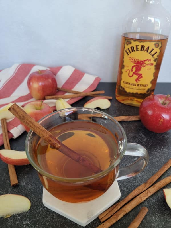 Apple cider fireball cocktail in a glass coffee mug with a cinnamon stick next to red apples, a cloth napkin, and bottle of fireball