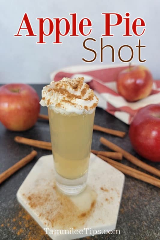 Apple Pie Shot over a shot garnished with whipped cream and cinnamon on a white coaster surrounded by cinnamon sticks and apples