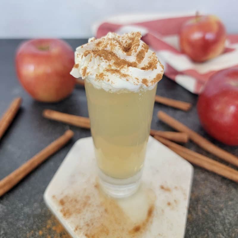 Apple pie shot garnished with whipped cream and cinnamon next to cinnamon sticks and red apples