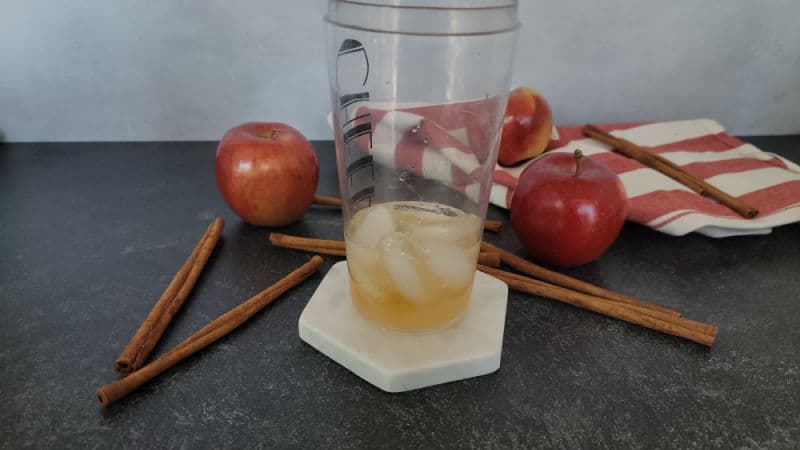 ice and liquid in a cocktail shaker next to cinnamon sticks and apples