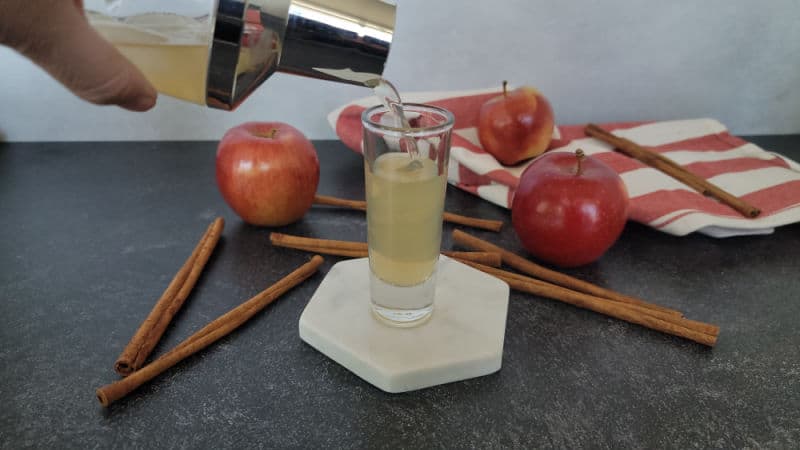 Cocktail shaker pouring an apple pie shot into a shot glass on a white coaster next to cinnamon sticks and apples
