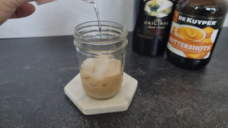 Clear buttershots pouring over Irish Cream in a mason jar glass on a marble coaster