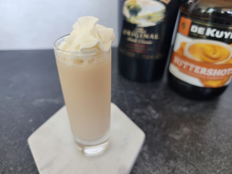 Buttery nipple shot with whipped cream garnish next to a bottle of Baileys and Butterscotch Schnapps