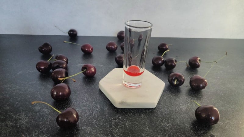 red liquid in a glass shot glass surrounded by fresh cherries 