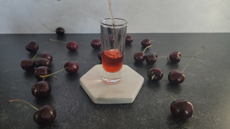 red liquid pouring into a shot glass on a white coaster surrounded by fresh cherries