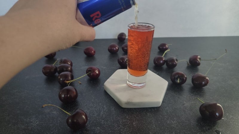 Red Bull pouring into a shot glass surrounded by fresh cherries