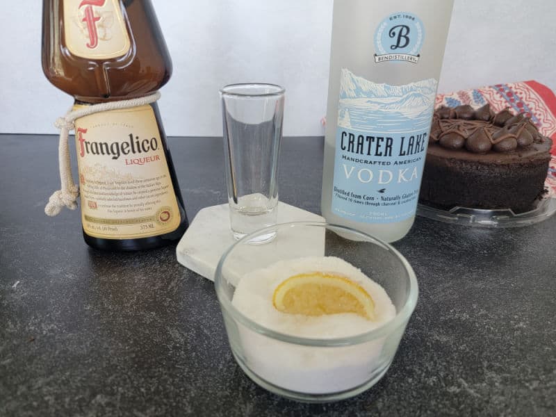 Frangelico, shot glass, bottle of vodka, lemon slice in a bowl of sugar next to a chocolate cake. 
