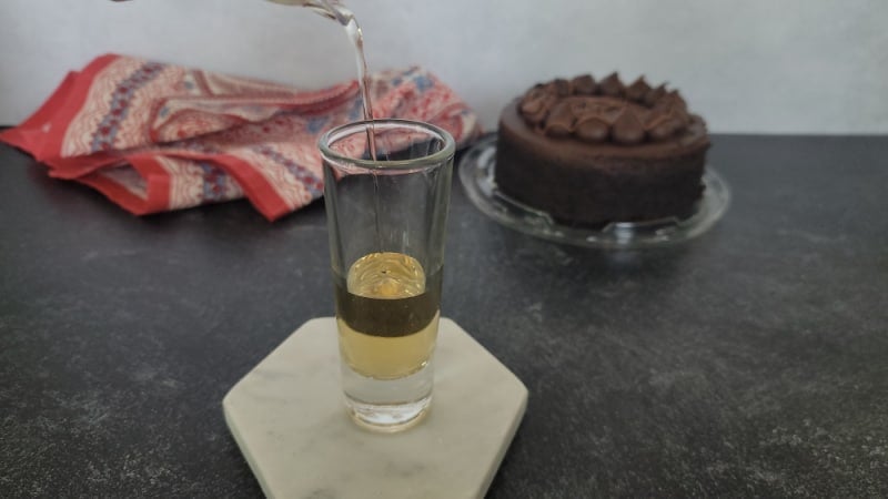 liquid pouring into a shot glass on a white coaster near a chocolate cake. 