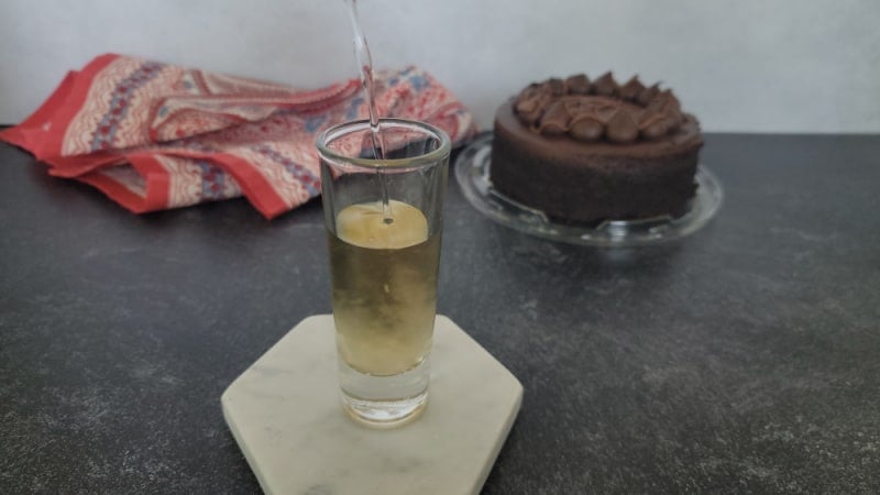Liquid pouring into a shot glass on a white coaster near a chocolate cake