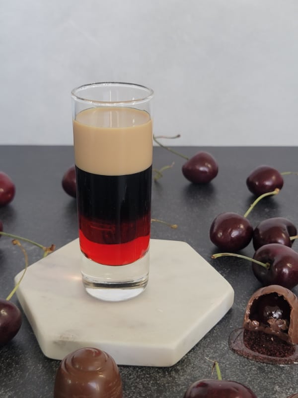 Chocolate covered cherry layered shot in a glass shot glass on a white coaster surrounded by chocolate covered cherry candies