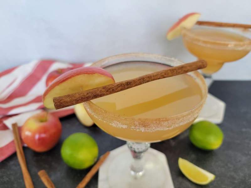 Apple cider margarita with a cinnamon stick and apple slice garnish next to red apples, limes and cinnamon sticks. 