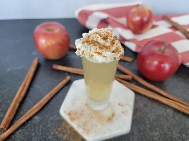 Apple pie shot garnished with whipped cream and cinnamon on a white coaster next to cinnamon sticks and red apples