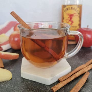 Fireball Apple Cider Recipe with cinnamon stick garnish in a clear glass coffee mug on a coaster next to apples and cinnamon sticks
