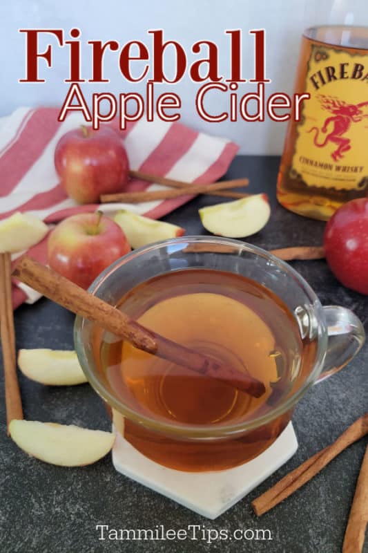 Fireball Apple Cider text over a glass with apple cider and a cinnamon stick next to a bottle of Fireball whisky, apples, and a red cloth napkin