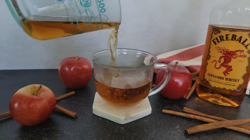 Hot apple cider pouring into a glass mug next to apples, cinnamon sticks, and a bottle of Fireball cinnamon whisky