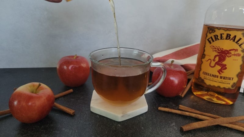 Liquid pouring into a coffee mug with apple cider next to red apples, cinnamon sticks, and a bottle of Fireball Cinnamon Whisky