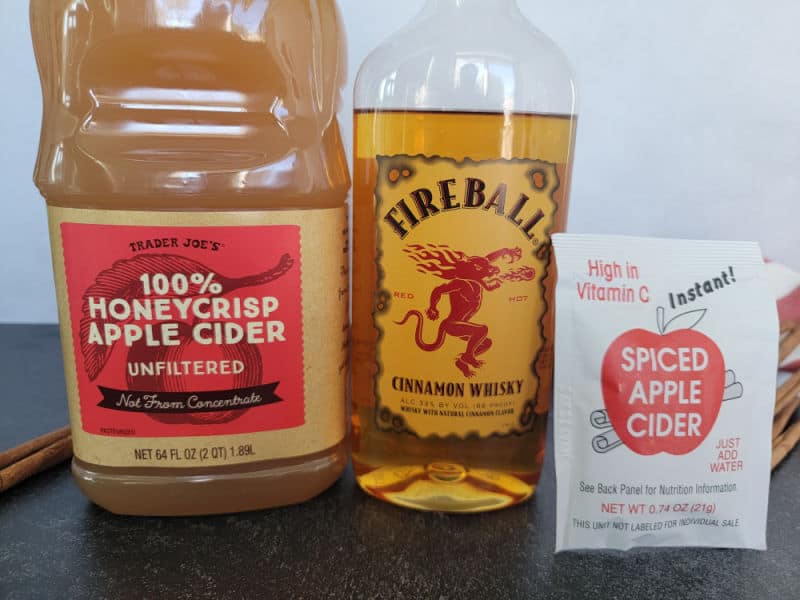 Trader joe's apple cider container, Fireball cinnamon whisky bottle, and spiced apple cider mix on a counter with cinnamon sticks. 