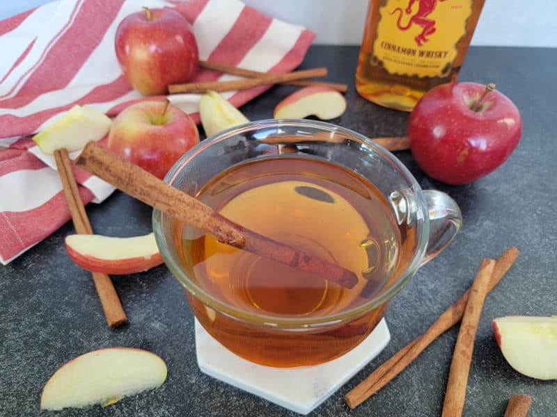 Fireball Apple Cider Cocktail in a glass coffee mug on a white coaster next to apple slices, cinnamon sticks, and a partial bottle of Fireball 