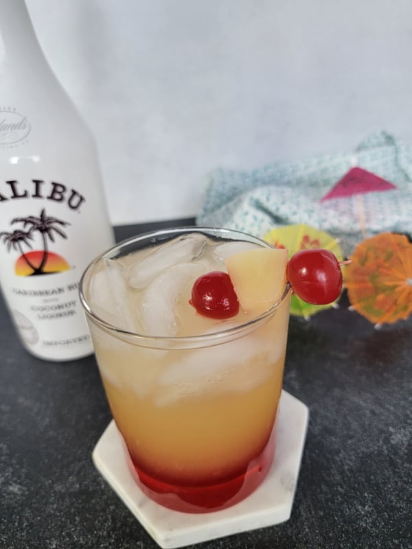 Layered yellow and red liquid in a clear glass with a garnish of maraschino cherries and pineapple chunks. Malibu coconut rum bottle in the background 