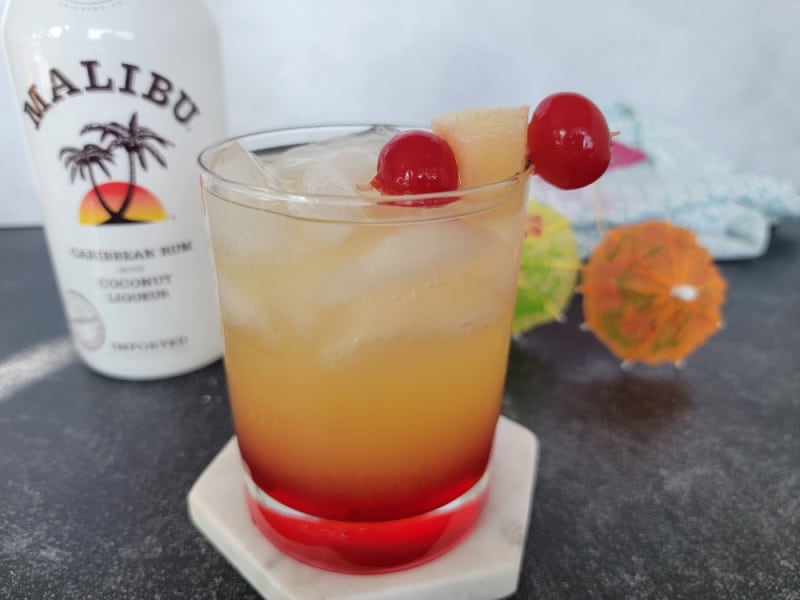 Red and yellow layered cocktail with maraschino cherry and pineapple chunk garnish in front of a bottle of Malibu coconut rum. 