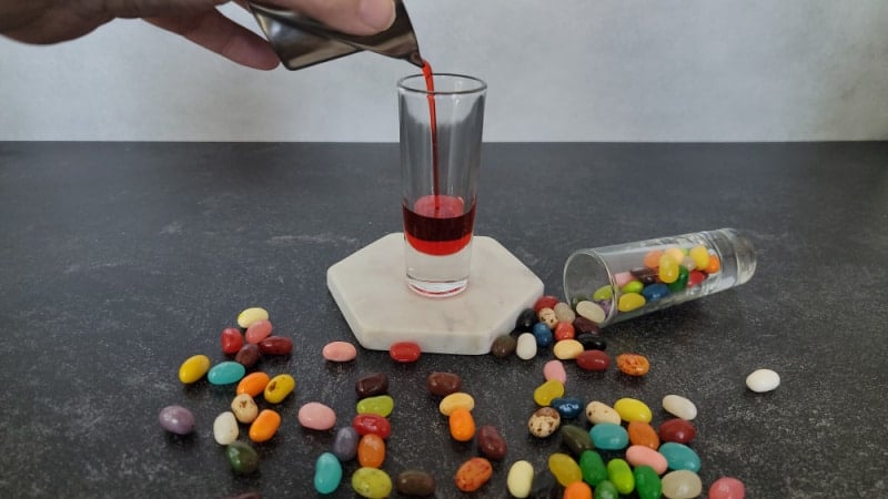 Red liquid pouring into a shot glass on a white coaster surrounded by jelly beans