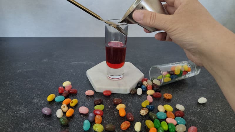 bar spoon in a shot glass above red liquid with a jigger about to pour into the glass. Jelly beans spilled across the counter. 