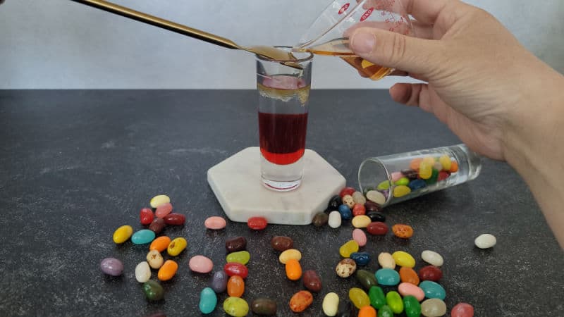 gold bar spoon in a shot glass next to a hand holding a jigger about to pour into the glass. Jelly beans spilled across in front of the shot glass