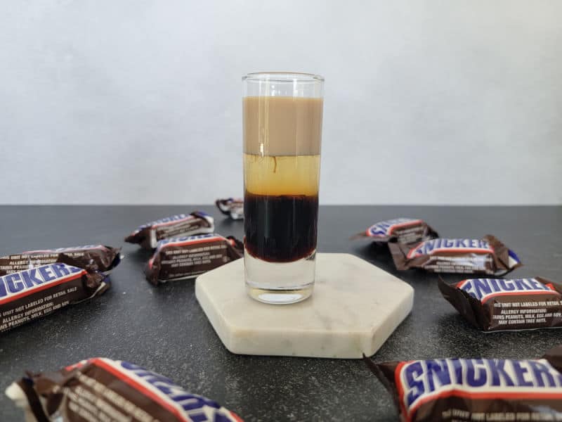 Layered Snickers Shot on a white coaster surrounded by Snickers Chocolate Bars