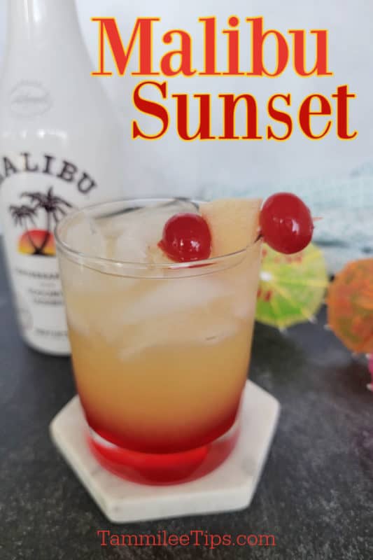 Malibu Sunset over a glass with orange and red liquid, garnishes with cherries and a pineapple chunk. In front of a bottle of Malibu Rum and tropical umbrellas