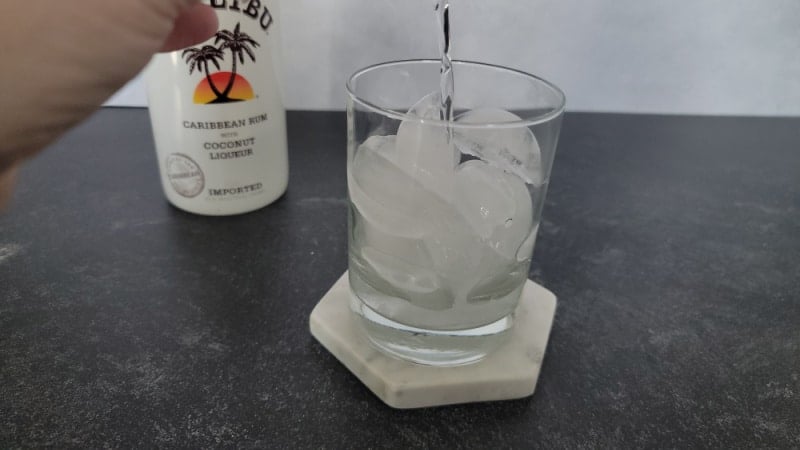 Clear liquid poured into a glass with ice cubes. Bottle of Malibu in the background. 