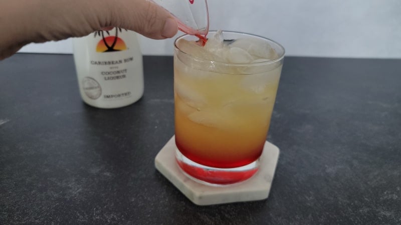 Red liquid pouring into a yellow liquid in a clear glass with a bottle of Malibu rum in the background. 
