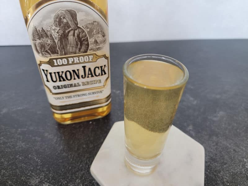 Snakebite shot in a shot glass on a white coaster next to a bottle of Yukon Jack