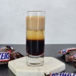 Layered Snickers Shot on a white coaster surrounded by Snickers Chocolate Bars