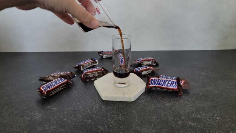 Kahlua pouring into a shot glass with Snickers Candy Bars nearby