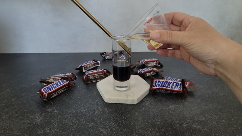 Frangelico pouring over the Kahlua in a shot glass with Snickers Candy Bars nearby
