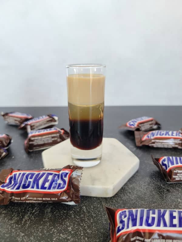 Layered Snickers Shot on a white coaster surrounded by Snickers Chocolate Bars