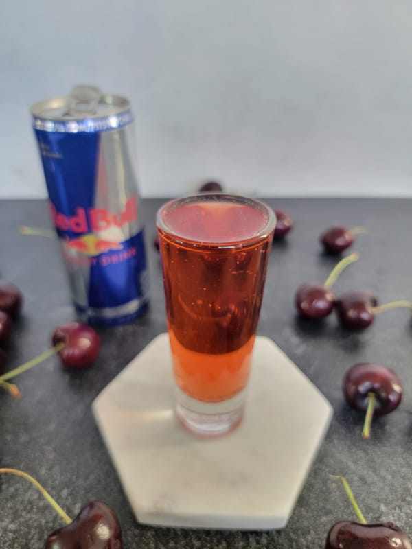 red cocktail shot on a white coaster with a can of Red Bull and Fresh cherries near it. 