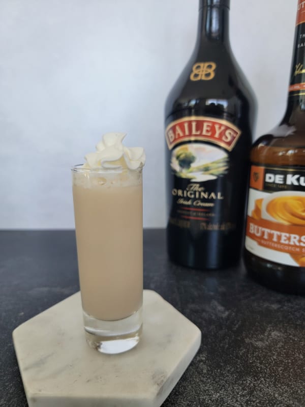 Buttery Nipple Shot garnished with whipped cream in a tall shot glass on a marble coaster with bottles in the background