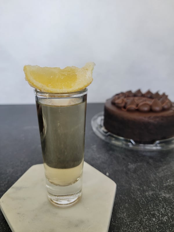 sugar covered lemon sitting on a shot glass on a white coaster next to a chocolate cake. 