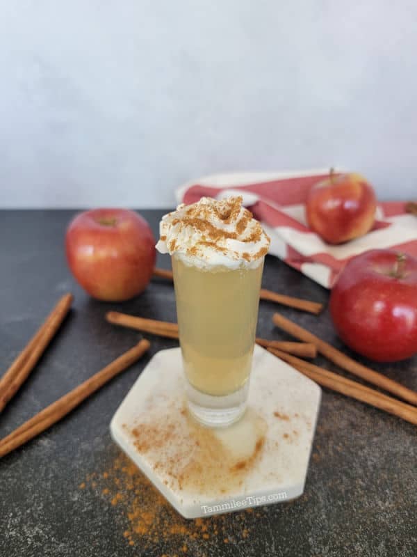 Apple pie shot with whipped cream and cinnamon next to cinnamon sticks and red apples