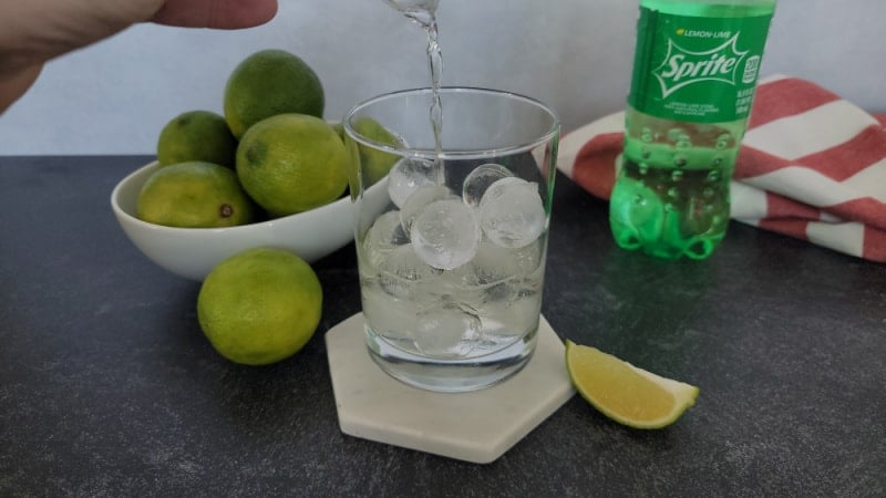 rum pouring into a cocktail glass next to a bowl of limes and a bottle of Sprite